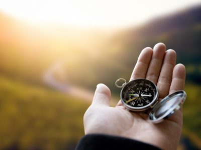 Traveler explorer man holding compass in a hand in mountains at sunrise, point of view.